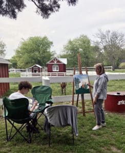 A lady and young man paint with easels while looking at a meadow and small barn