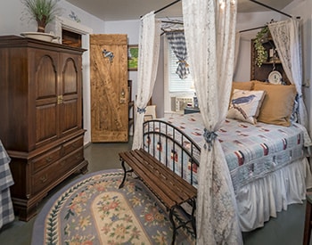 A light bluebonnet colored room with white lace drapes on bed and wooden furniture and blue rug with wooden door to room