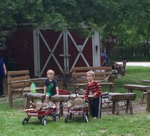 2 young boys hold red wagons that say Firewood on them by a stone firepit