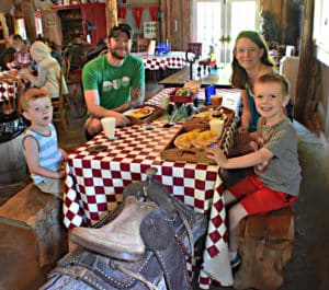 A family of four sits around a checkered table in a barn eating breakfast