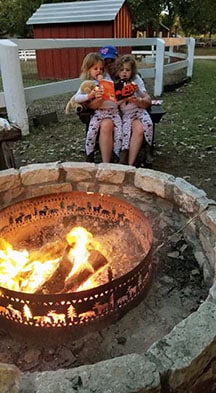 A dad holds two young twin daughters in his lap reading a book to them by a campfire.