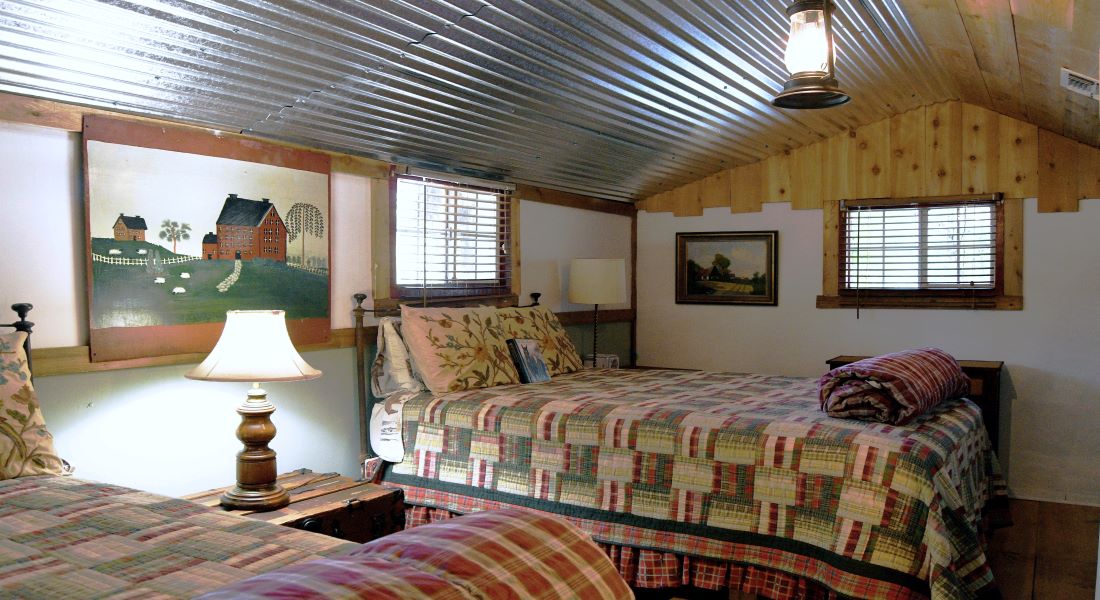 Two beds with red and green and cream squares with a trunk and lamp between them, windows, tin ceiling and wooden trim