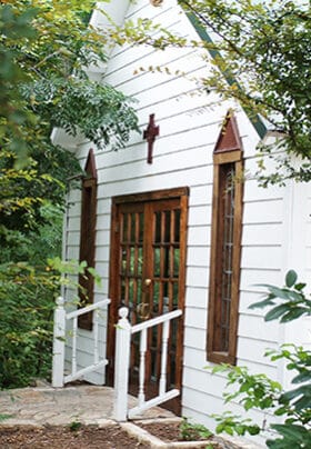 The front of a white wooden chapel with brown wooden window trim in a wooded area with green trees and stone path