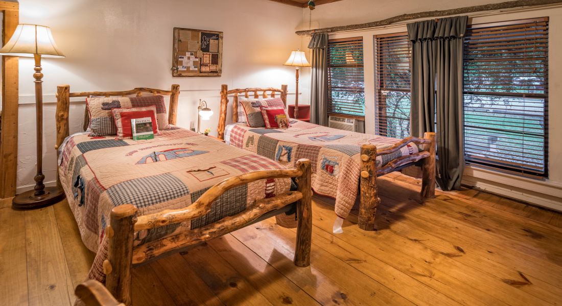 Two log cabin style twin beds with camping theme quilts with a wooden plank floor and large floor to ceiling windows along one wall
