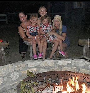 Three moms and 2 young twin girls sit in laps around a campfire.