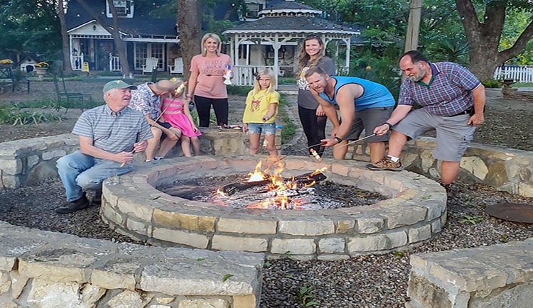 A family of all ages roasts marshmallows around a stone campfire pit
