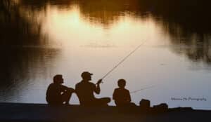 An evening silhouette of 3 men with fishing rods sitting on a dam with the river behind them
