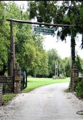 An entrance to a green lawn with a cement road going under a wooden sign saying Country Woods Inn as a gated entrance