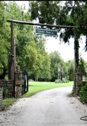 An entrance to a green lawn with a cement road going under a wooden sign saying Country Woods Inn as a gated entrance