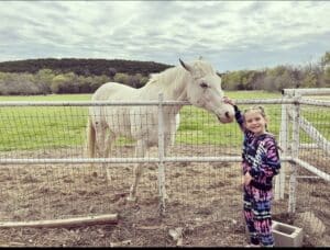 A young girl with blonde hair pets a white horse in front of a large meadow of wildflowers