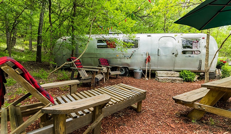 A silver Airstream sits in the woods with wooden adirondack chairs and picnic table surroundings