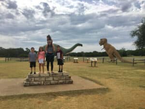 A mom with 3 kids stands on a stone base in front of two large dinosaur statues