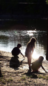 Silouhuettes of 3 kids squatting and standing by a river with the sun shining off of it