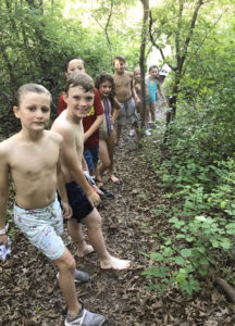 A group of young kids stand in a line along a walking trail in the woods in shorts with walking sticks