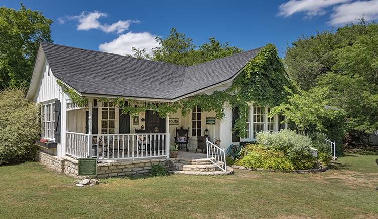 A white farm house with green ivy growing along the porch, campfire pit out front and black rocking chairs on the porch