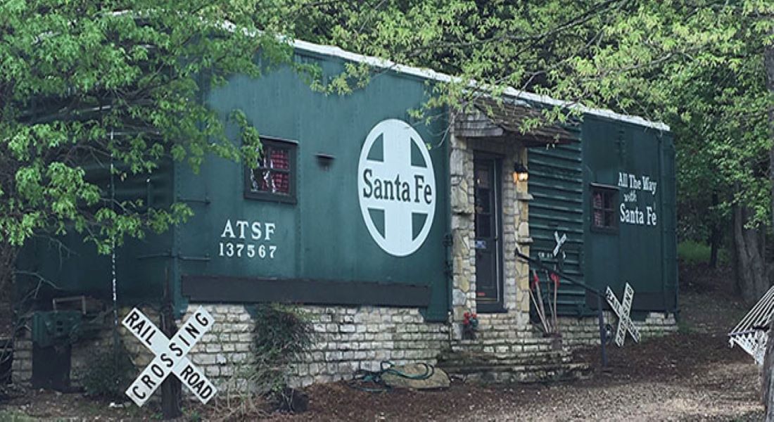 A dark green Santa Fe Rail Car sits on top of a stone foundation with stone steps to the door in a wooded area with a driveway and trees