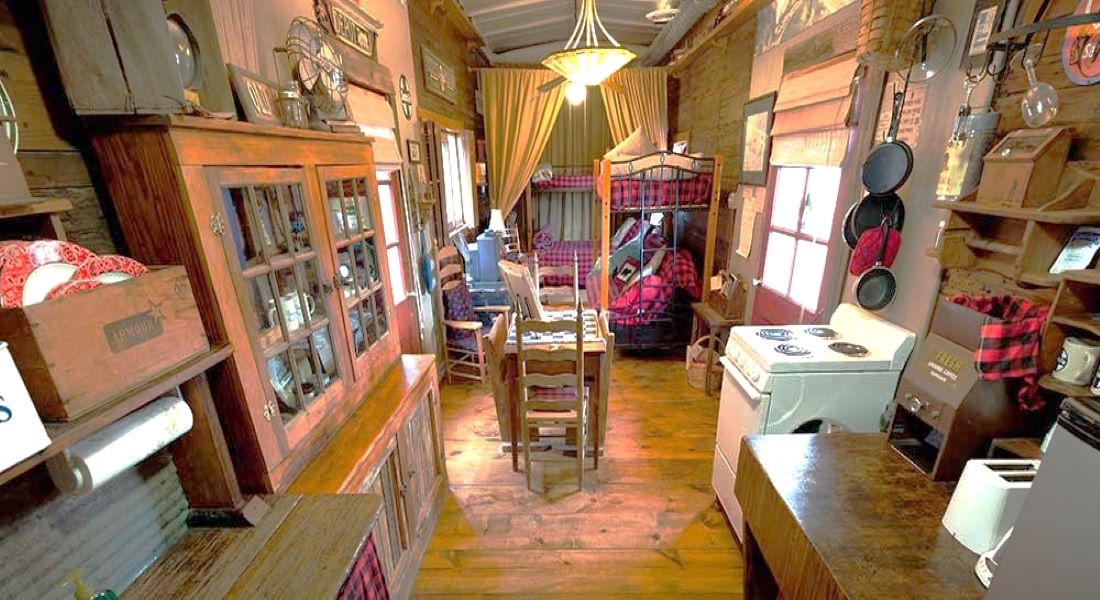 A long angle picture of a white stove on the right with two sets of twin bunk beds in the background with red and black check quilts with a stone floor