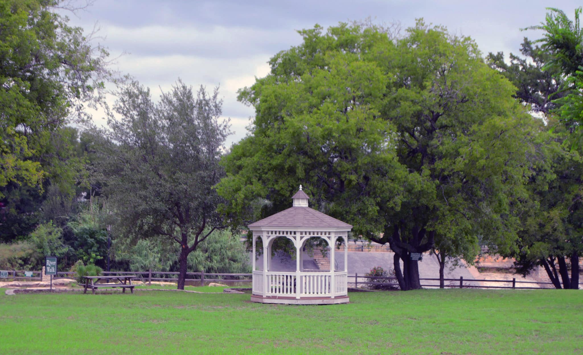 A white gazebo sits in a green meadow in front of a river