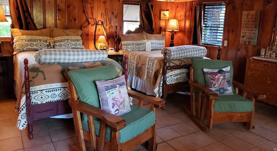 Two green cloth chairs with wooden frame sit on a saltillo tile floor with 2 full size beds behind them