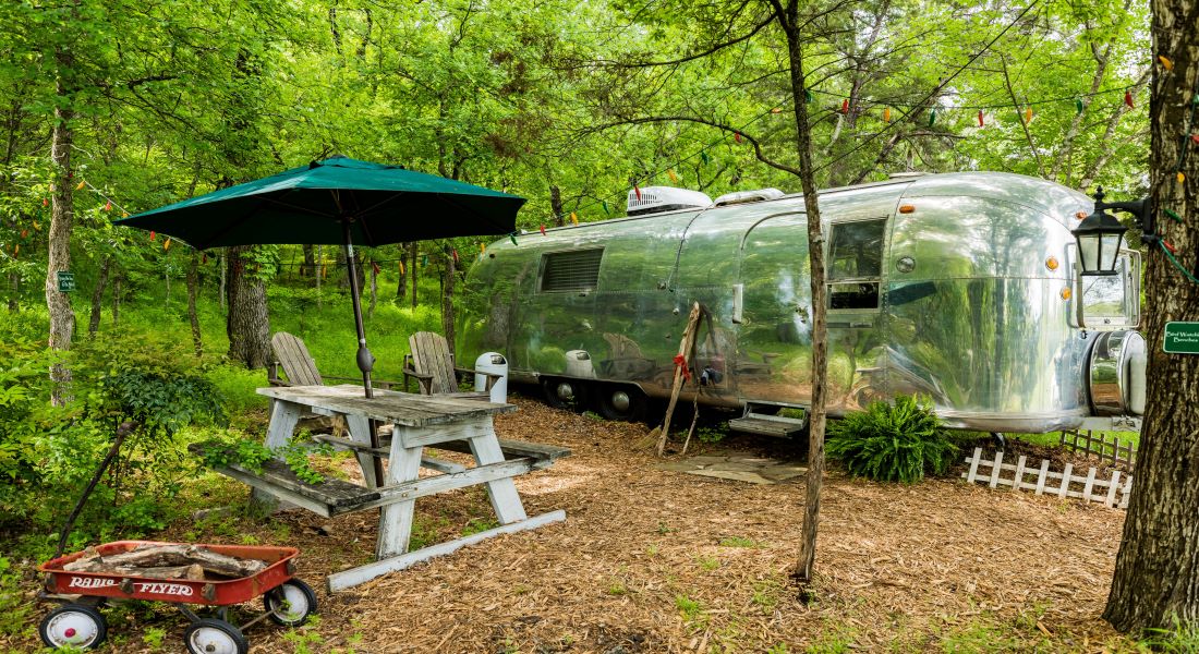 A silver Airstream trailer sits in a wooded area surrounded by green trees with mulch picnic area and wooden picnic table