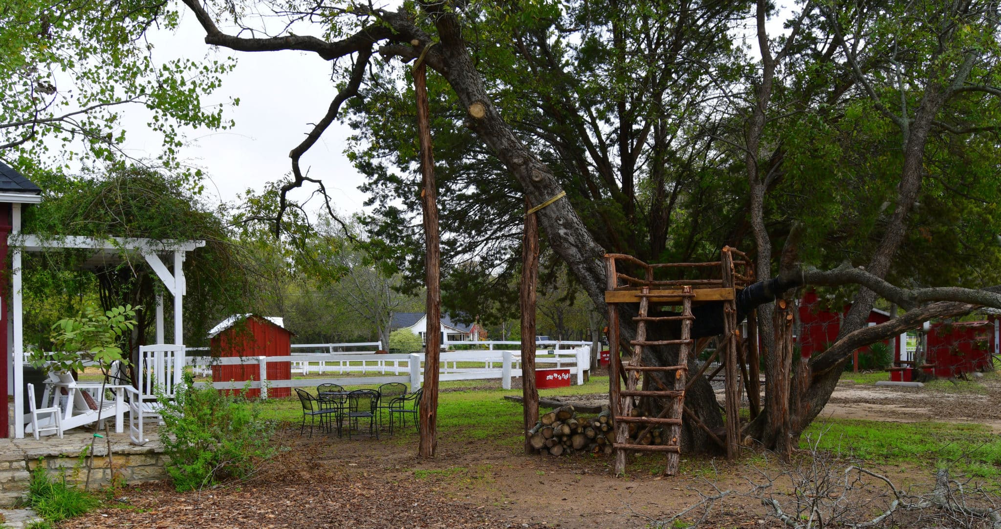 A cedar tree house stands in front of a white corral and red barn