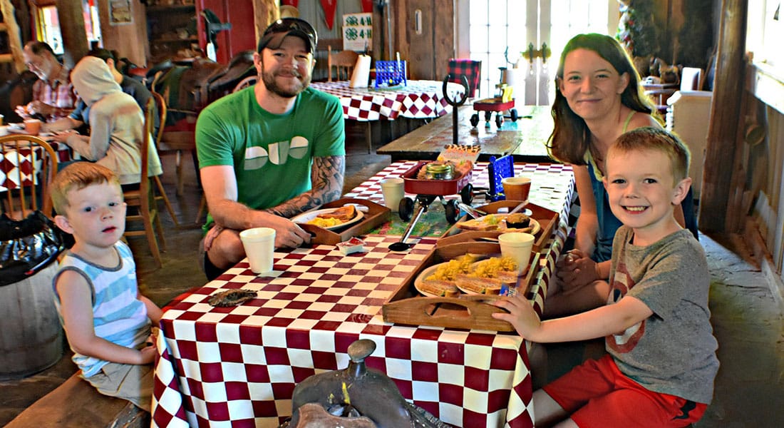 A mom, dad and two sons sit around a red and white checkered table in the breakfast barn eating eggs with a saddle for a seat.