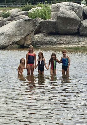 5 kids in swimsuits stand knee deep in the river water in bathing suits.