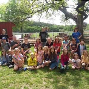 Large group of elementary aged kids sitting in a group on the lawn of a farm