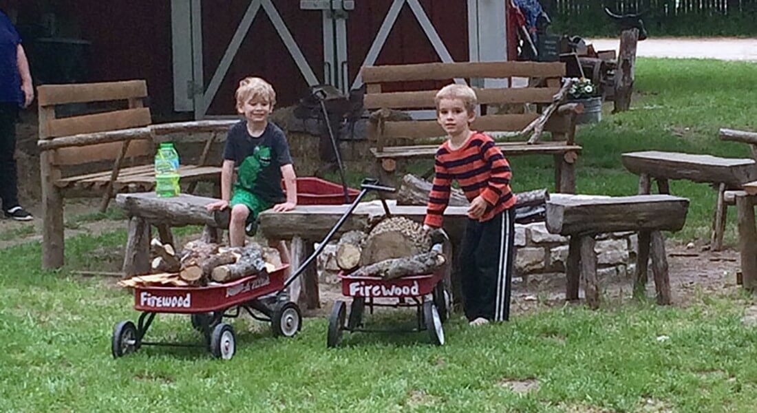 Two little blonde boys site on wooden benches neat red wagons full of firewood. 