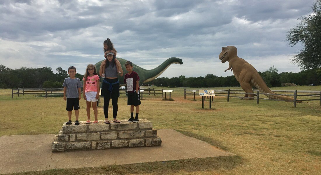 A group of kids stands on a stone plinth in a dinosaur park. 