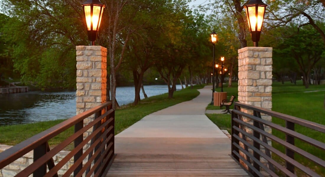 A path lit by carriage lamps along a river at dusk. 