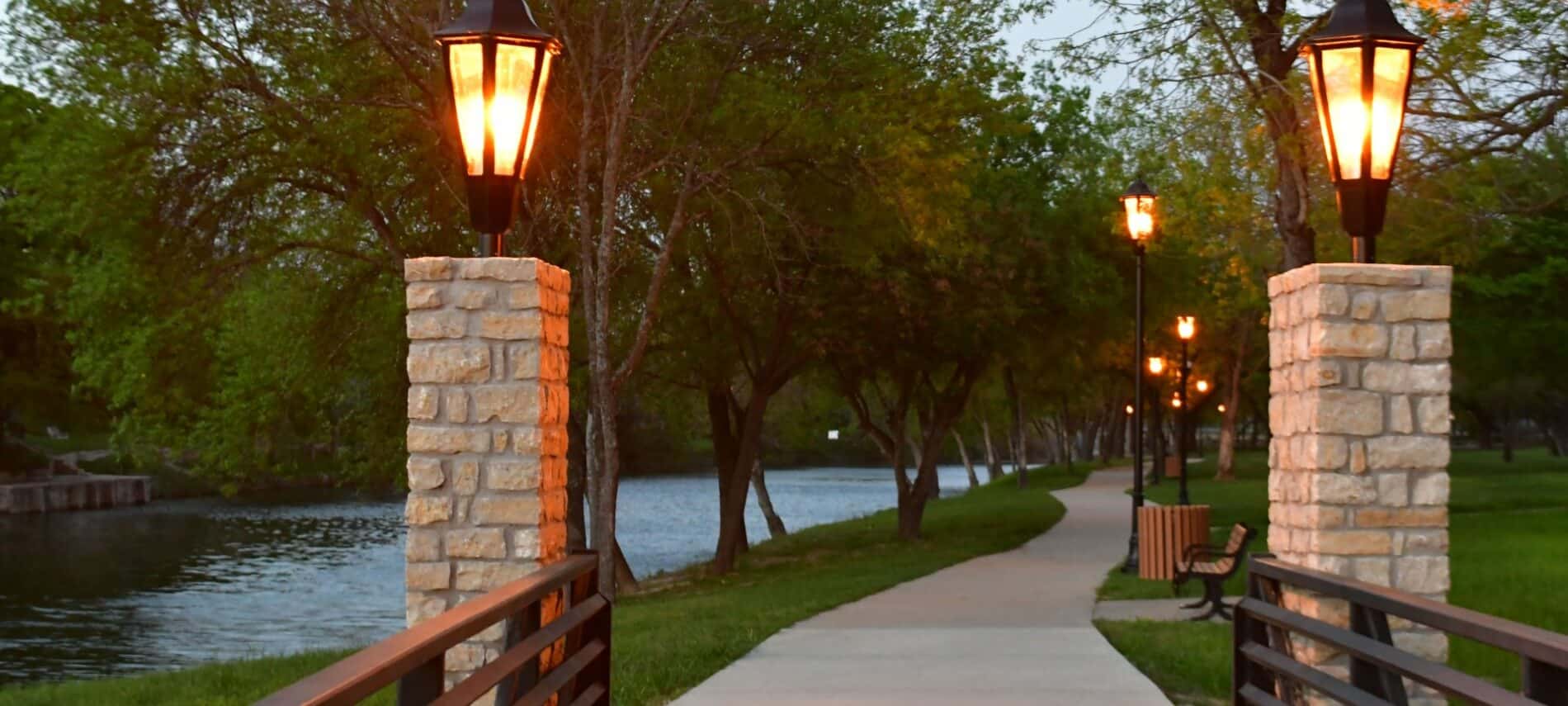 A path lit by carriage lamps along a river at dusk.