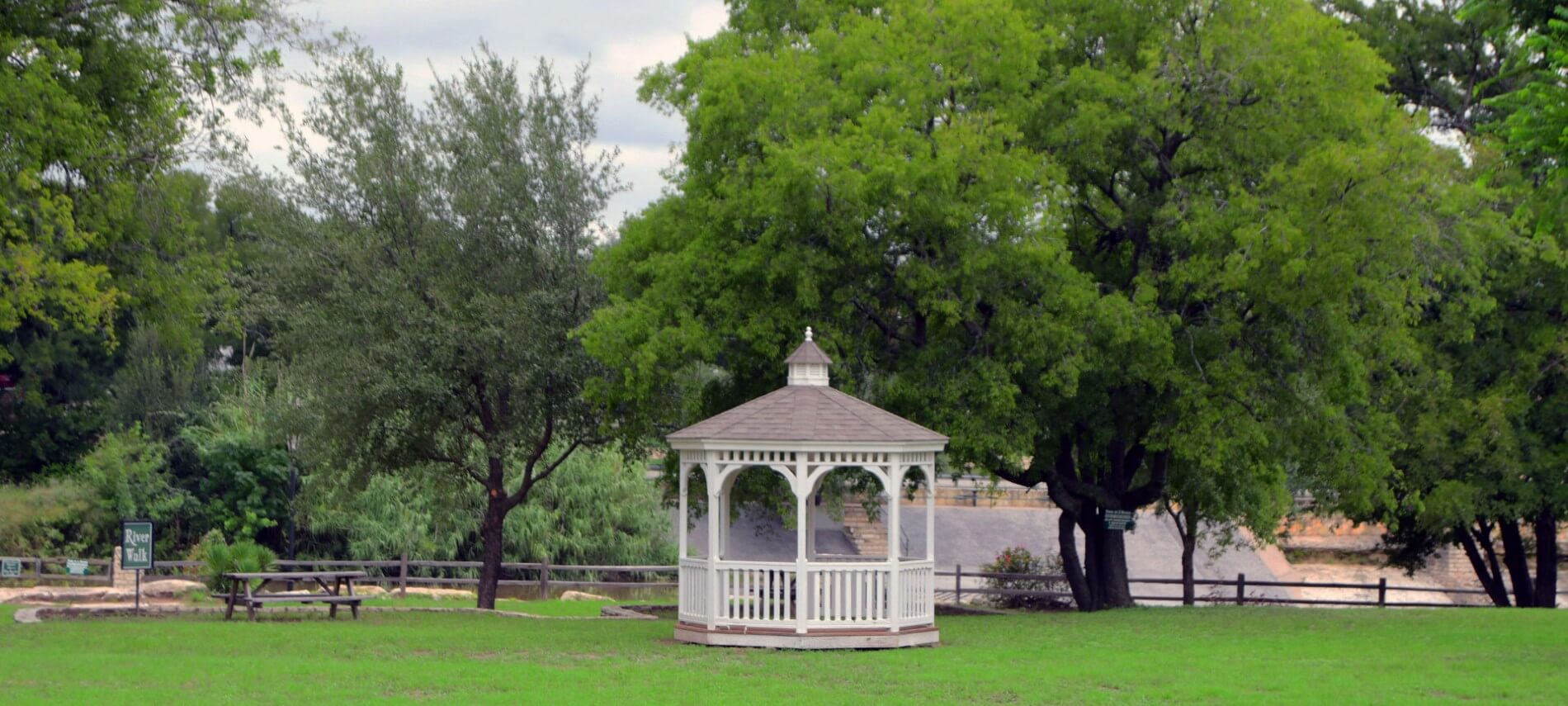 A pretty white gazebo in a green field surrounded by trees.