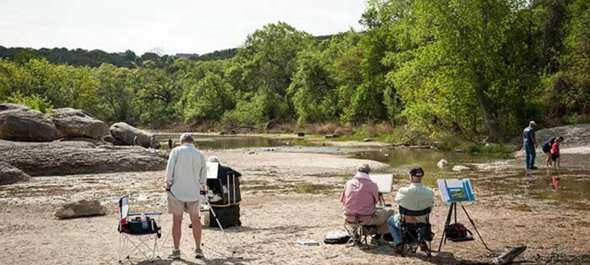 People paint, wade and sit next to a stream surrounded by green trees.