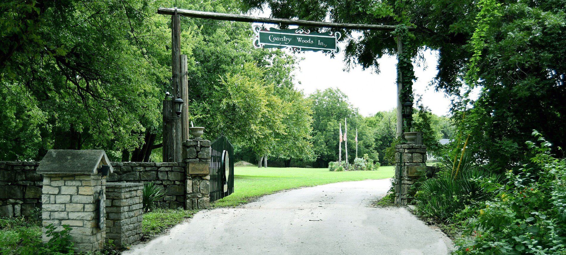 A country road surrounded by green trees leading to a stone entrance with a sign on a tall overhead wooden post.