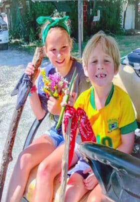 A young blonde boy and girl hold walking sticks with bandanas and wildflowers they have picked