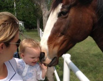 A blonde woman holds a baby close to a chesnut horse with a white blaze.