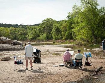 People paint, wade and sit next to a stream surrounded by green trees.