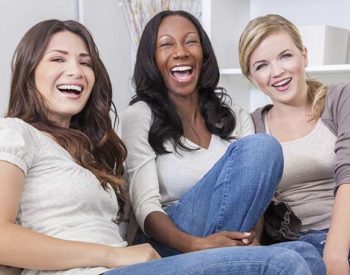 Three young ladies in jeans laugh together on a sofa.